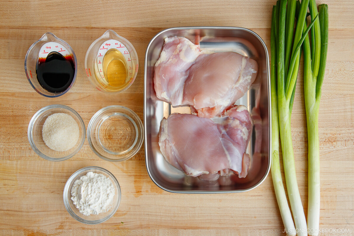 Raw chicken thighs on a metal tray, green onions, soy sauce, sake, sugar, flour, and cornstarch in glass bowls—all ingredients neatly arranged for a delicious yakitori don—set out on a wooden surface.