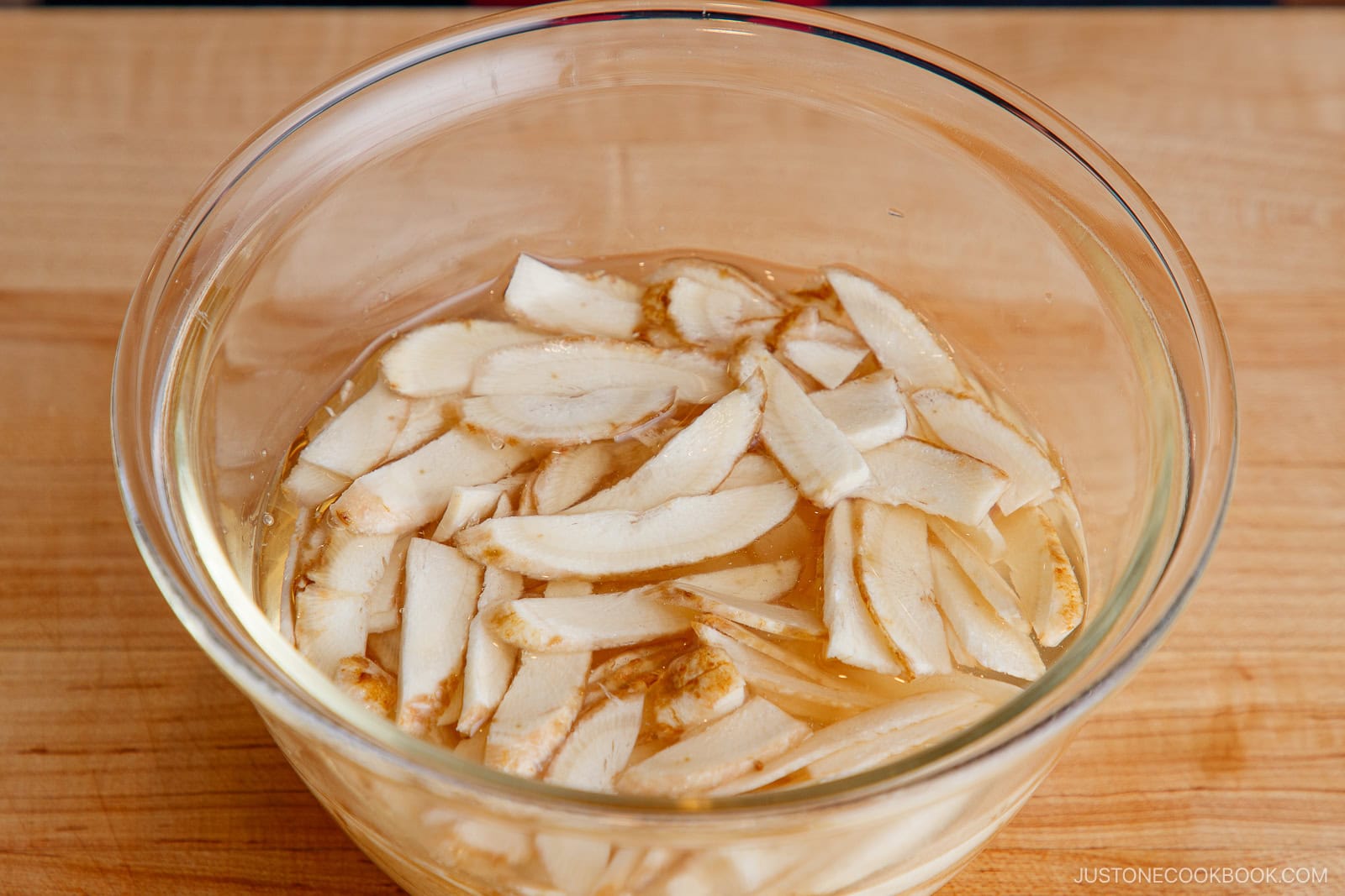 A glass bowl filled with thinly sliced burdock root soaking in water, ready to be used for a savory Beef and Gobo Stir Fry, sits atop a wooden surface.