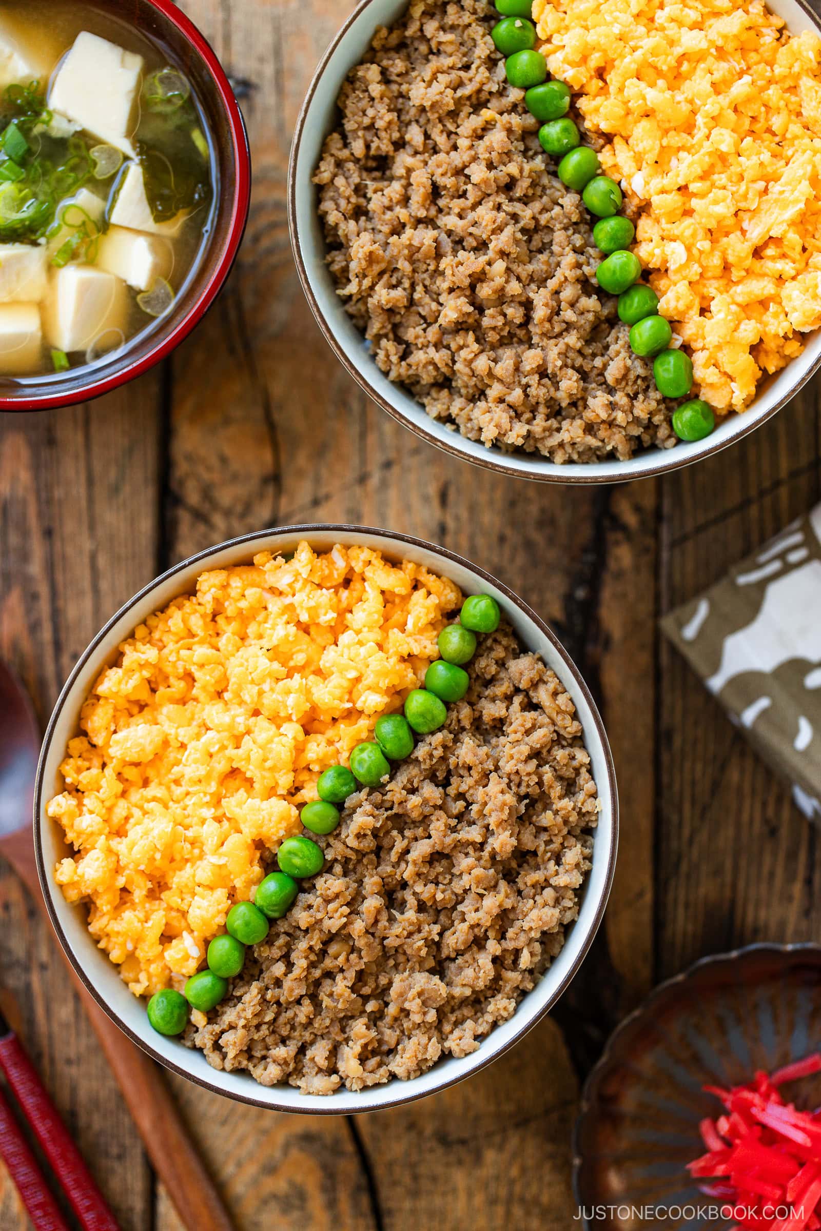 Two bowls of soboro don (ground chicken bowl) with seasoned meat, scrambled eggs, and green peas in neat sections. A bowl of miso soup with tofu and green onions sits beside them on a rustic wooden table.