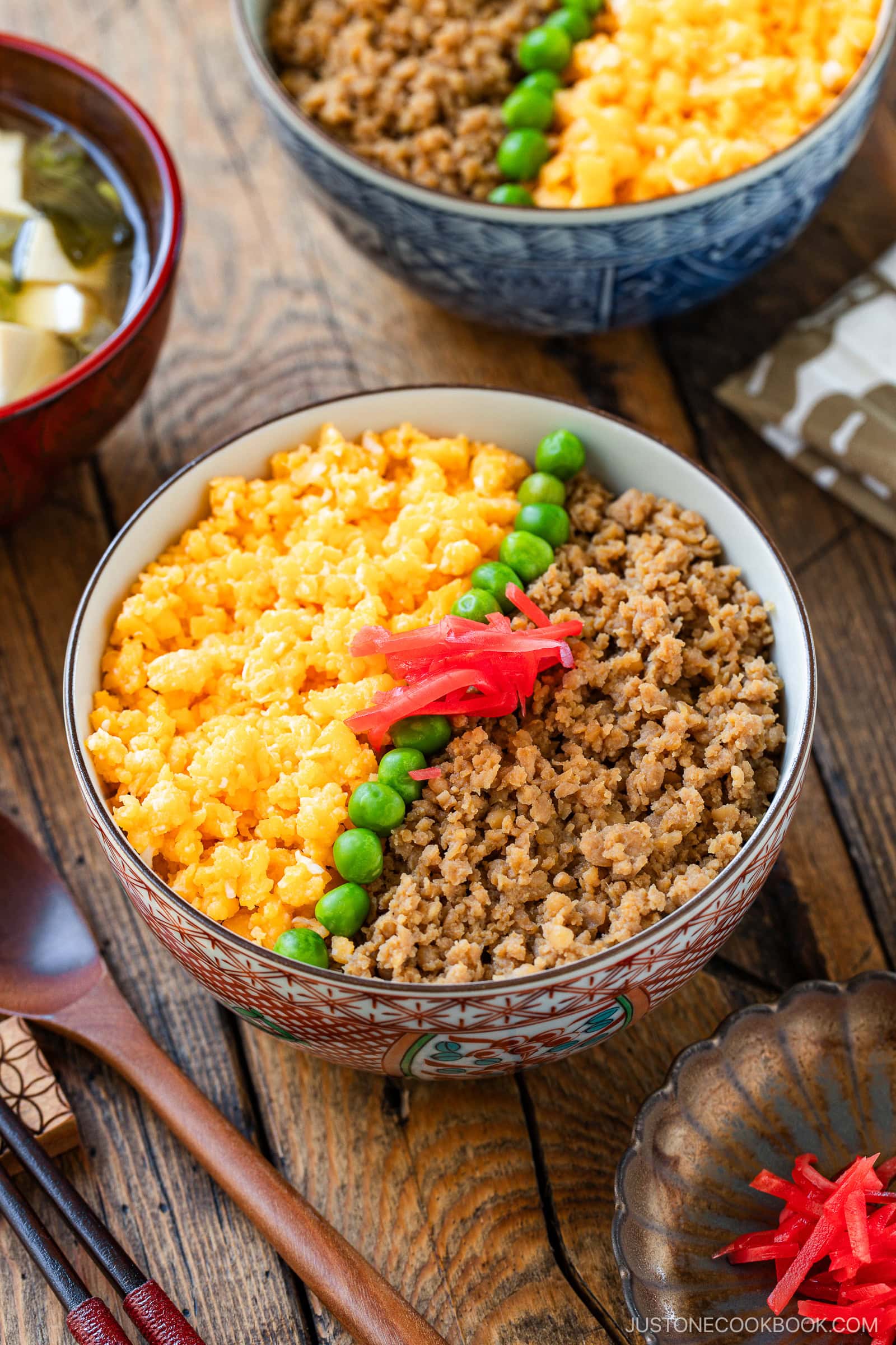 A bowl of soboro don (ground chicken bowl) with scrambled eggs, green peas, and pickled ginger arranged in sections, served on a wooden table with a bowl of miso soup and chopsticks beside it.