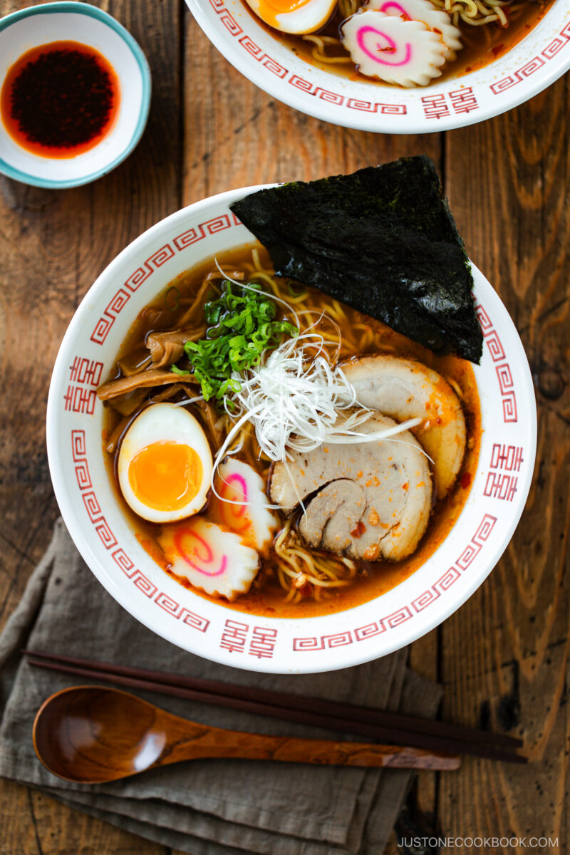 A bowl of spicy shoyu ramen with sliced pork, half a soft-boiled egg, narutomaki, seaweed, green onions, and white shredded garnish in a red and white patterned bowl on a wooden table. A spoon and chopsticks rest nearby.