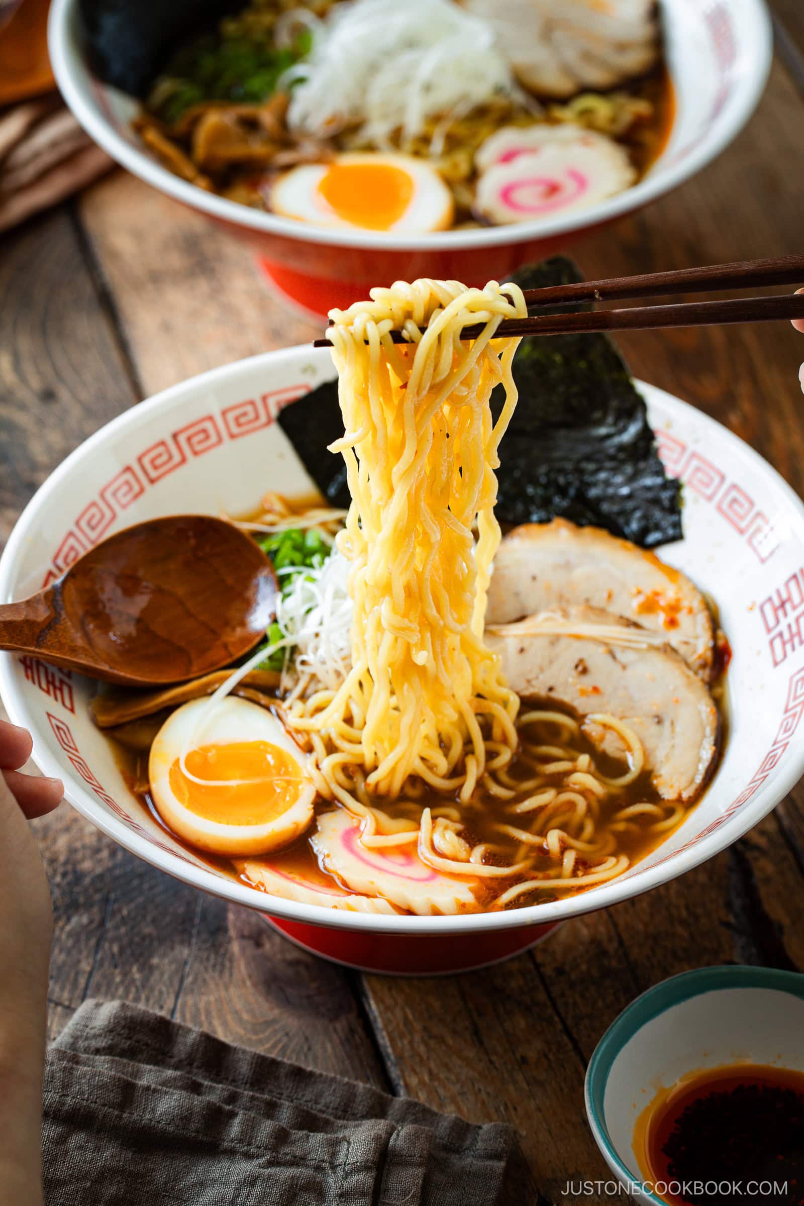 A hand uses chopsticks to lift yellow ramen noodles from a bowl of spicy shoyu ramen. The bowl contains sliced pork, a soft-boiled egg, narutomaki, seaweed, green onions, and broth, with a wooden spoon on the side.