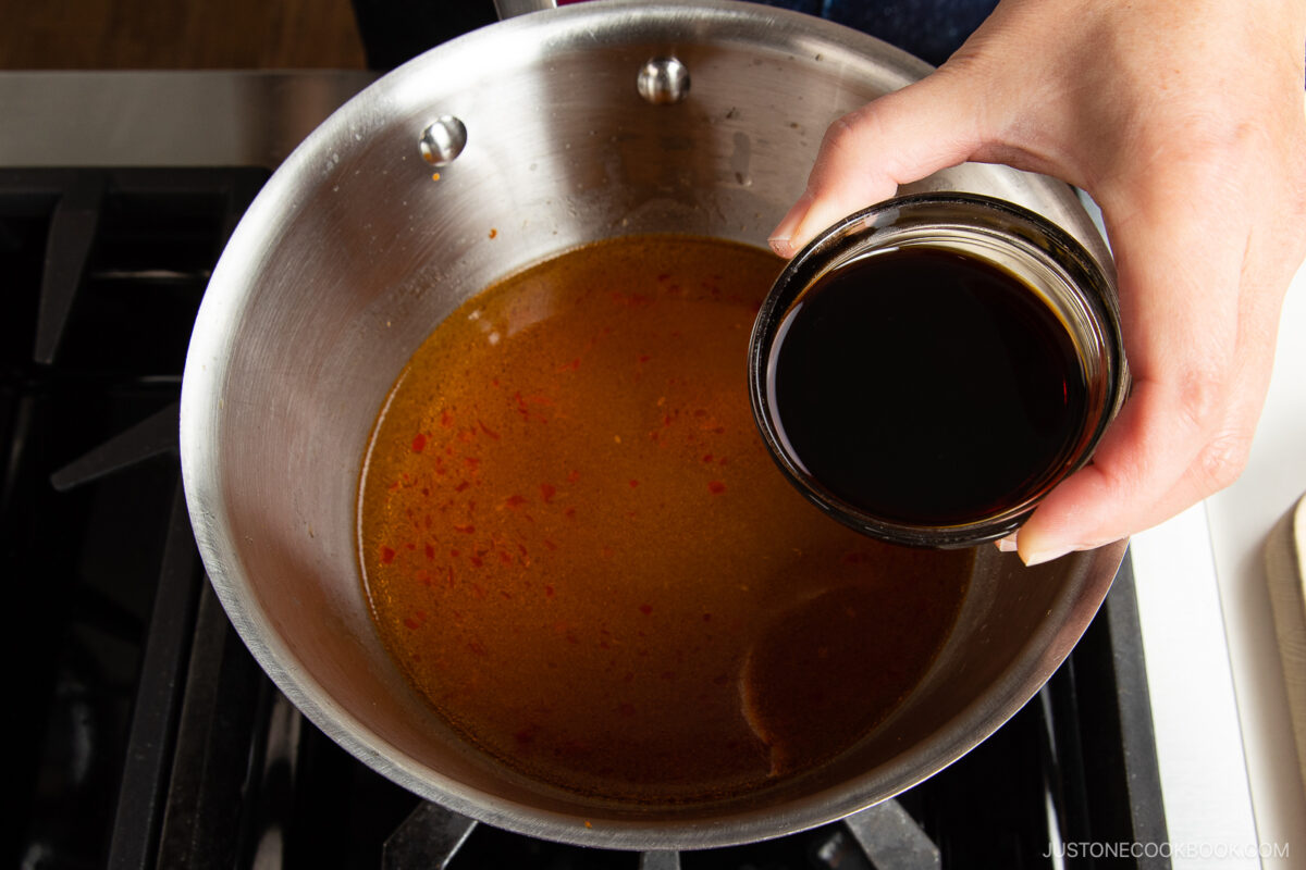 A hand holds a small bowl of dark liquid above a saucepan filled with reddish-brown broth, as if about to pour it in—capturing the moment of preparing spicy shoyu ramen on the stovetop.