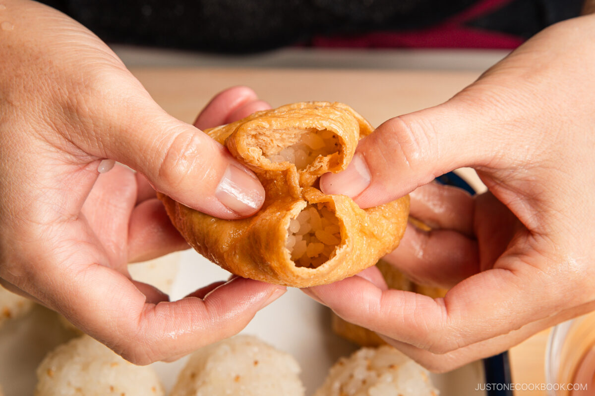 Two hands pull open a piece of inari sushi, revealing rice nestled inside the tofu skin pouch. Several more inari sushi rice balls are visible in the background on a plate.