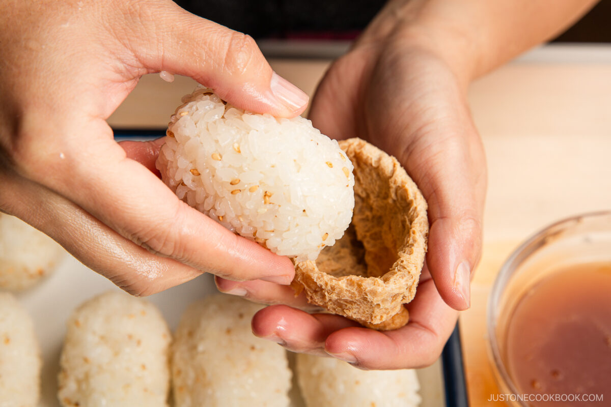 A person stuffs a ball of sticky rice with sesame seeds into an open pouch of seasoned tofu skin, preparing inari sushi. Several rice balls and a bowl of sauce are visible nearby.