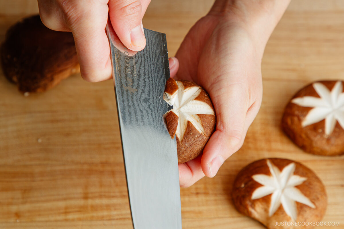 A hand demonstrates how to make a flower pattern on shiitake mushroom caps, using a knife to score a star-shaped design, with other beautifully scored mushrooms resting on a wooden cutting board nearby.