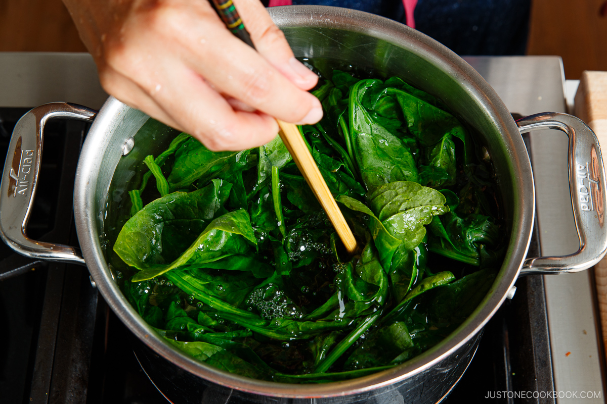 A hand uses chopsticks to stir fresh spinach leaves boiling in a stainless steel pot on a stove, showing essential prep work for a delicious meal.