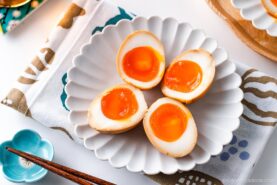 Four ramen eggs, halved to reveal their soft, runny yolks, are arranged on a white scalloped plate set on a patterned cloth. A pair of wooden chopsticks and a blue dish complete this inviting ramen eggs presentation.