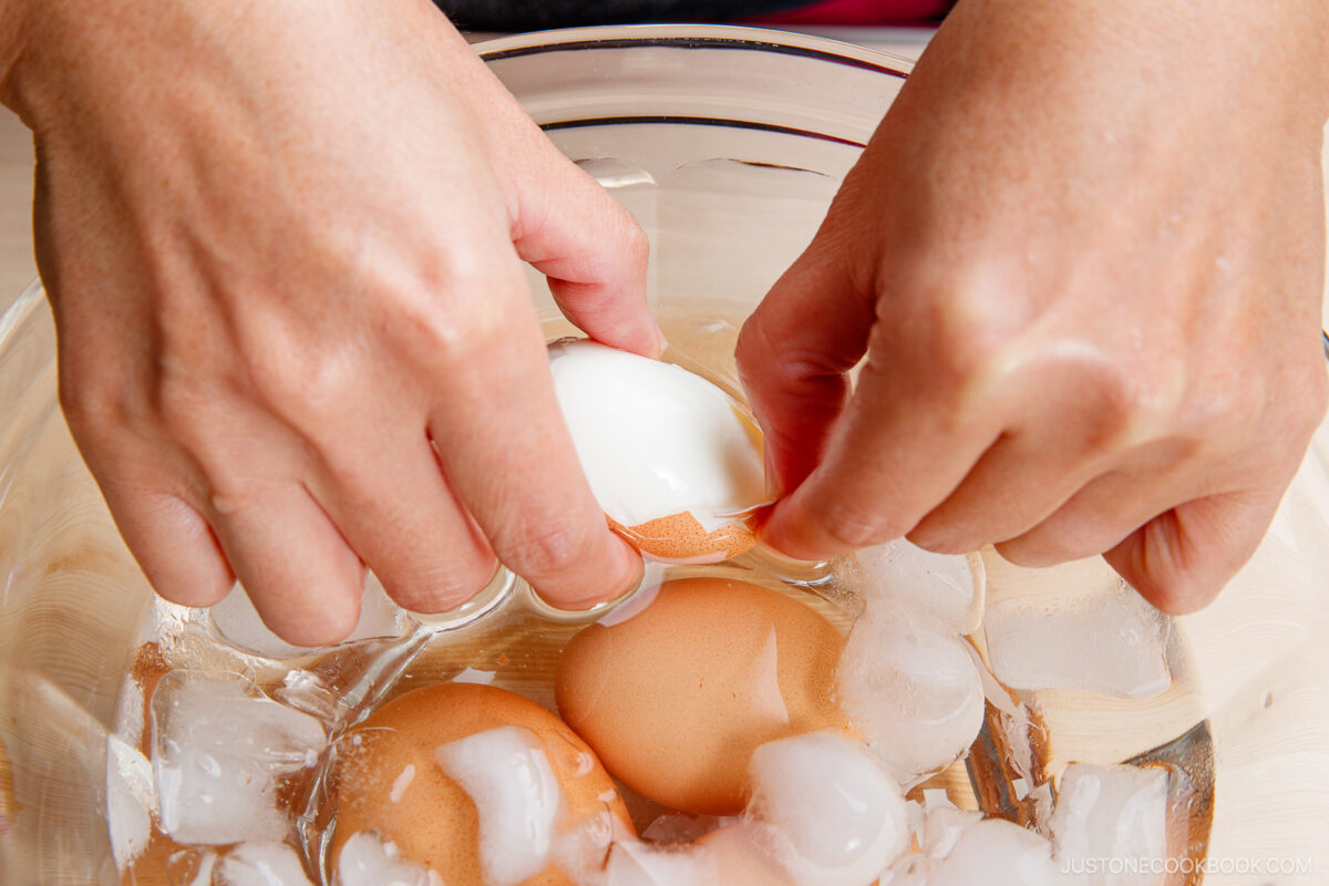 A person peeling the shell off a ramen egg over a bowl filled with ice water and other eggs.