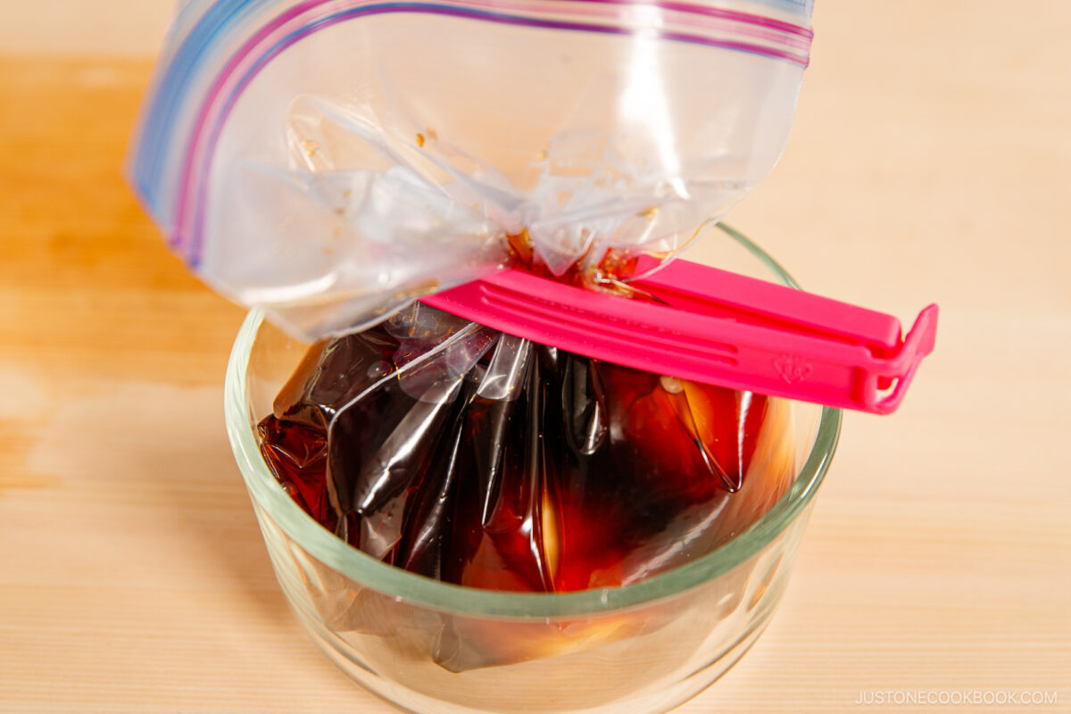 A clear plastic bag filled with dark marinade for ramen eggs is sealed with a pink clip and placed in a glass bowl on a wooden surface.