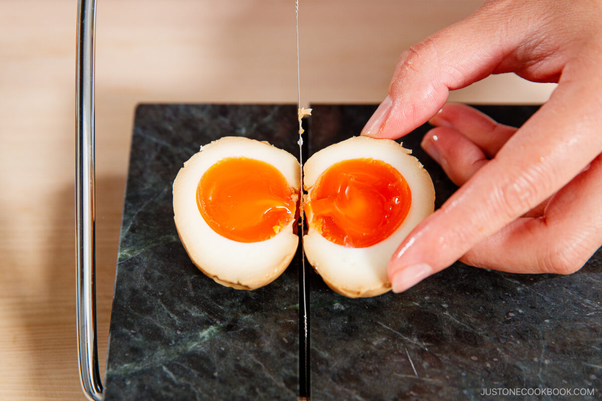 A hand slices a marinated ramen egg in half with a wire egg slicer, revealing its jammy orange yolk on a dark cutting board.