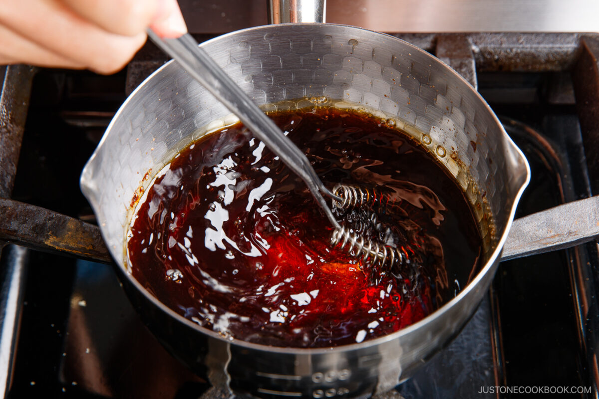 A hand stirs a dark, glossy liquid in a saucepan on a stovetop using a small whisk—perfect for preparing the flavorful marinade used to soak ramen eggs. The mixture simmers gently, its surface catching the light.