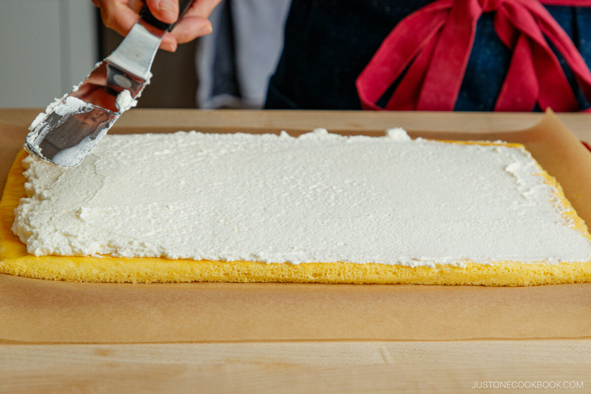 A person spreads a layer of white cream filling onto a rectangular yellow sponge cake, preparing a Japanese fruit roll cake on parchment paper set atop a wooden surface.
