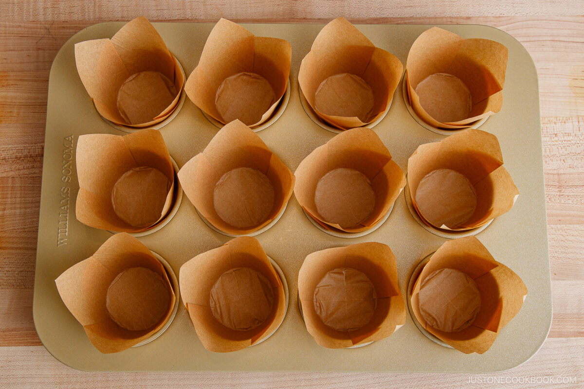 A gold muffin pan holds twelve empty brown parchment paper muffin liners, arranged neatly in each cup, set on a light wooden surface.