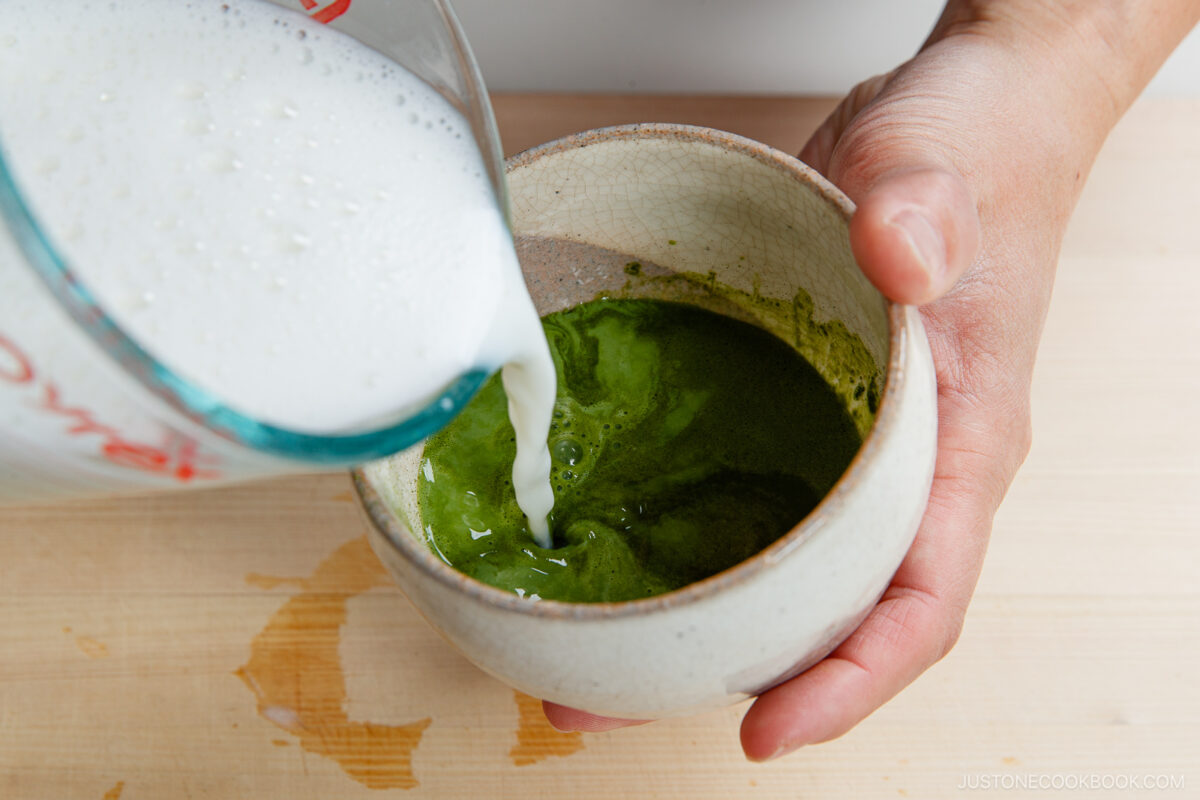 A hand holds a ceramic bowl of whisked green matcha as frothed milk is poured from a measuring cup, creating a vibrant matcha latte on a light wooden surface.