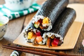 Two uncut sushi rolls—ehomaki (setsubun sushi roll)—filled with rice, vegetables, and seafood are wrapped in nori and arranged on a bamboo plate with chopsticks and a small teacup nearby.