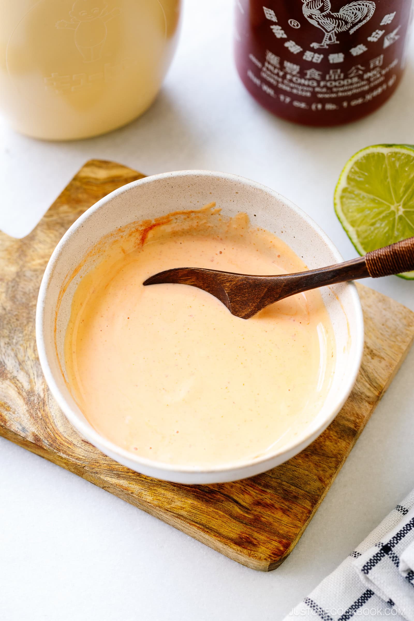 A bowl of creamy orange sauce, made from a homemade spicy mayo recipe, sits with a wooden spoon on a small board. Sriracha and mayonnaise bottles and a lime wedge are visible in the background.