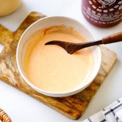 A bowl of creamy orange sauce—your homemade spicy mayo recipe—sits with a wooden spoon on a cutting board. Nearby are a halved lime, sriracha bottle, and checkered cloth on a white surface.