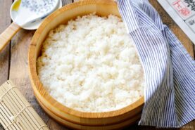 A wooden bowl filled with cooked white sushi rice, perfect for learning how to make sushi rice, sits on a wooden table, partially covered by a blue and white striped cloth with a bamboo sushi mat and ceramic spoon nearby.
