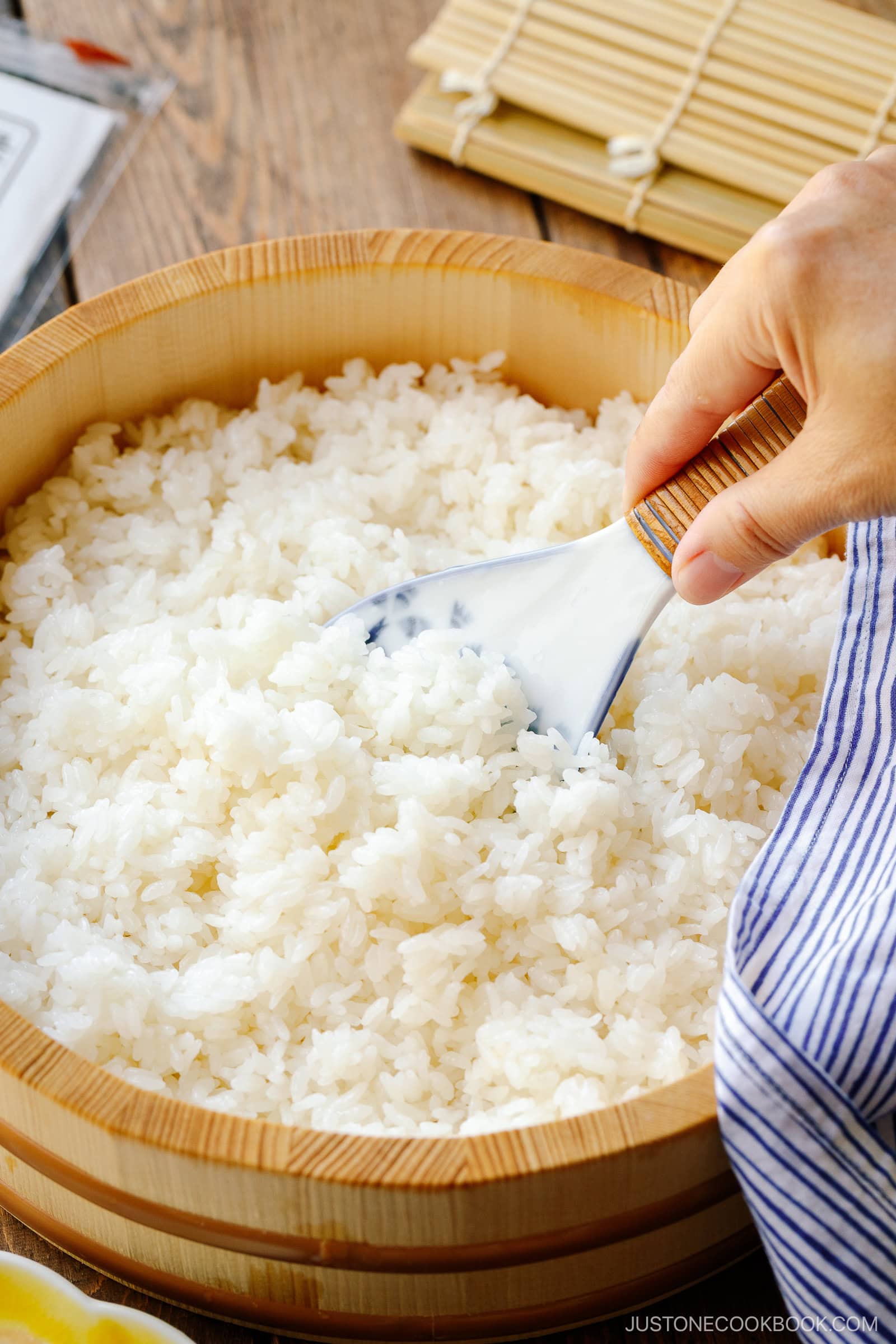 A hand demonstrates how to make sushi rice by mixing cooked rice with a paddle in a wooden bowl, while a bamboo sushi mat and striped cloth rest nearby on the wooden surface.