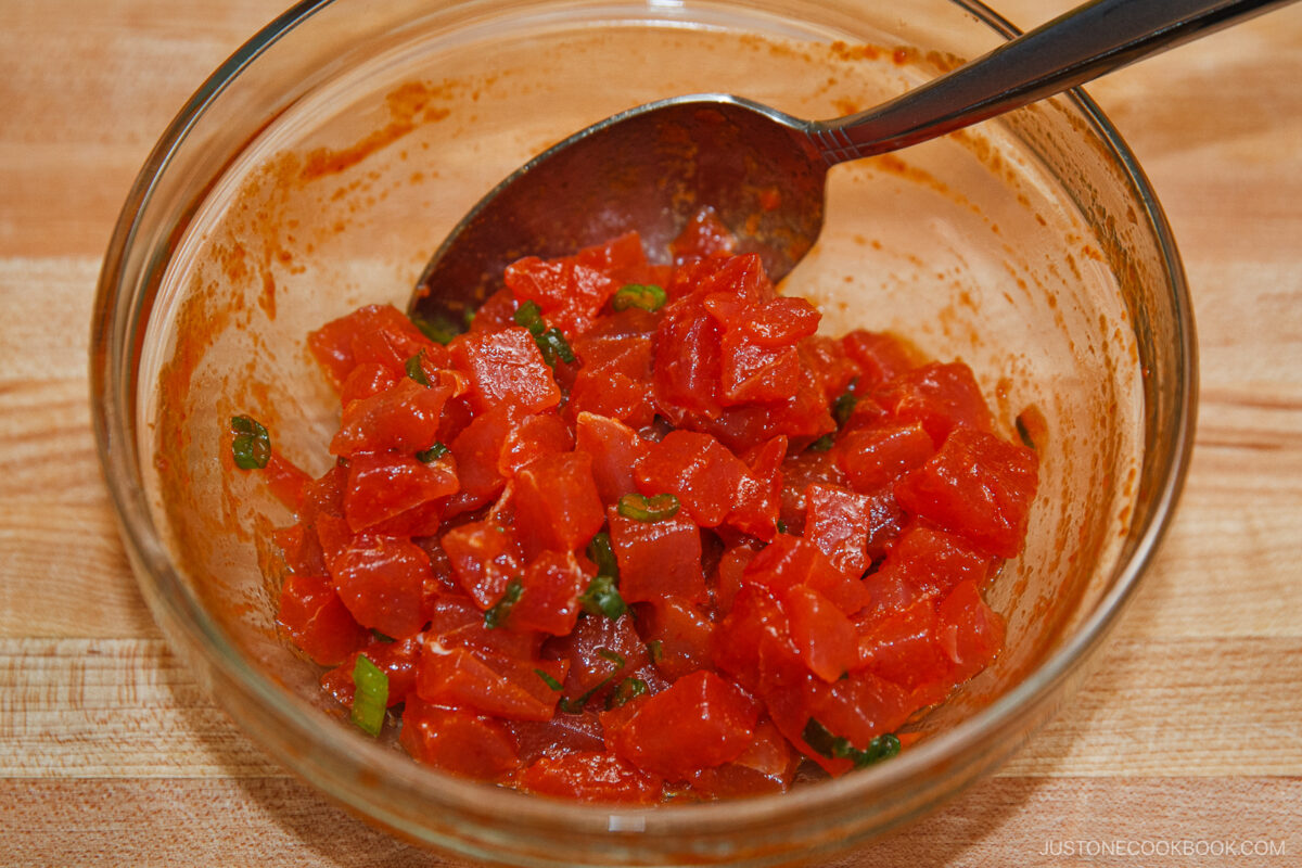 A glass bowl filled with diced raw tuna, green onions, and a reddish marinade—just like the filling for spicy tuna rolls—rests on a wooden surface with a metal spoon inside.