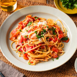 A plate of Tomato Bacon Pasta topped with fresh herbs and grated cheese sits on a woven placemat, with a glass of white wine, extra cheese, herbs, and red pepper flakes on the side. Silverware and a napkin are nearby.