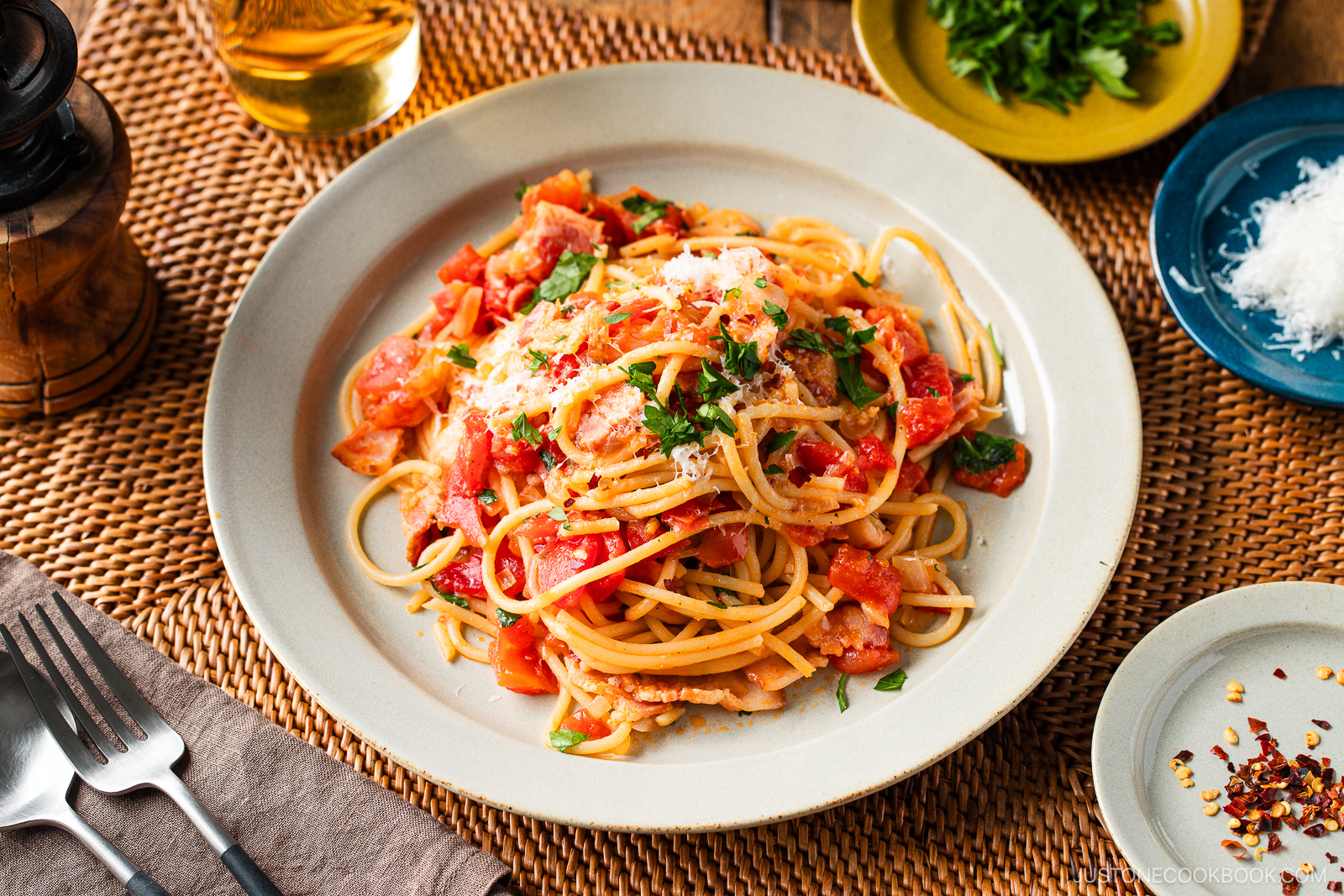A plate of Tomato Bacon Pasta topped with fresh herbs and grated cheese sits on a woven placemat, with a glass of white wine, extra cheese, herbs, and red pepper flakes on the side. Silverware and a napkin are nearby.