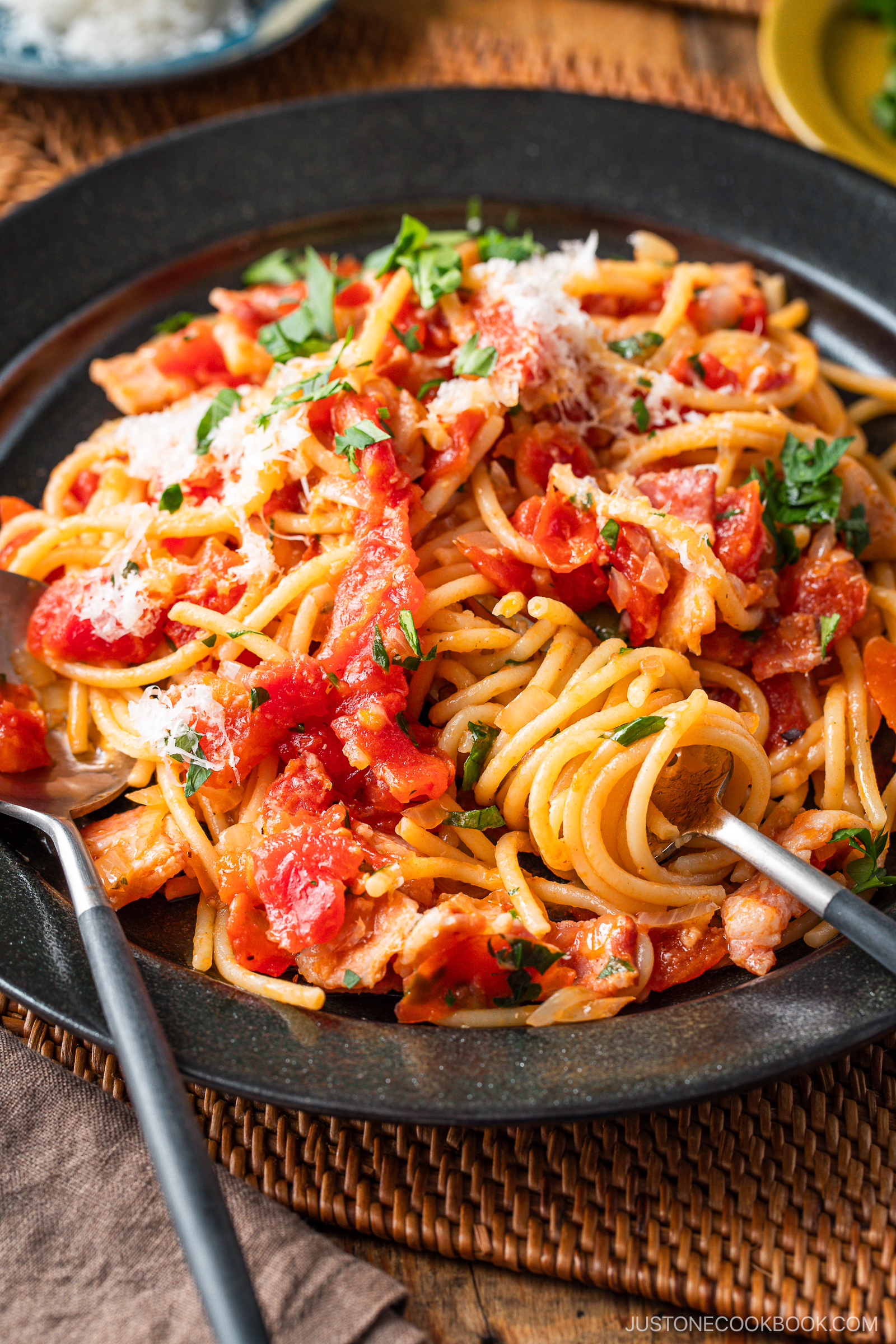 A plate of Tomato Bacon Pasta topped with tomato sauce, chunks of tomato, crispy bacon, grated cheese, and chopped parsley, served with a fork and spoon on a dark plate.