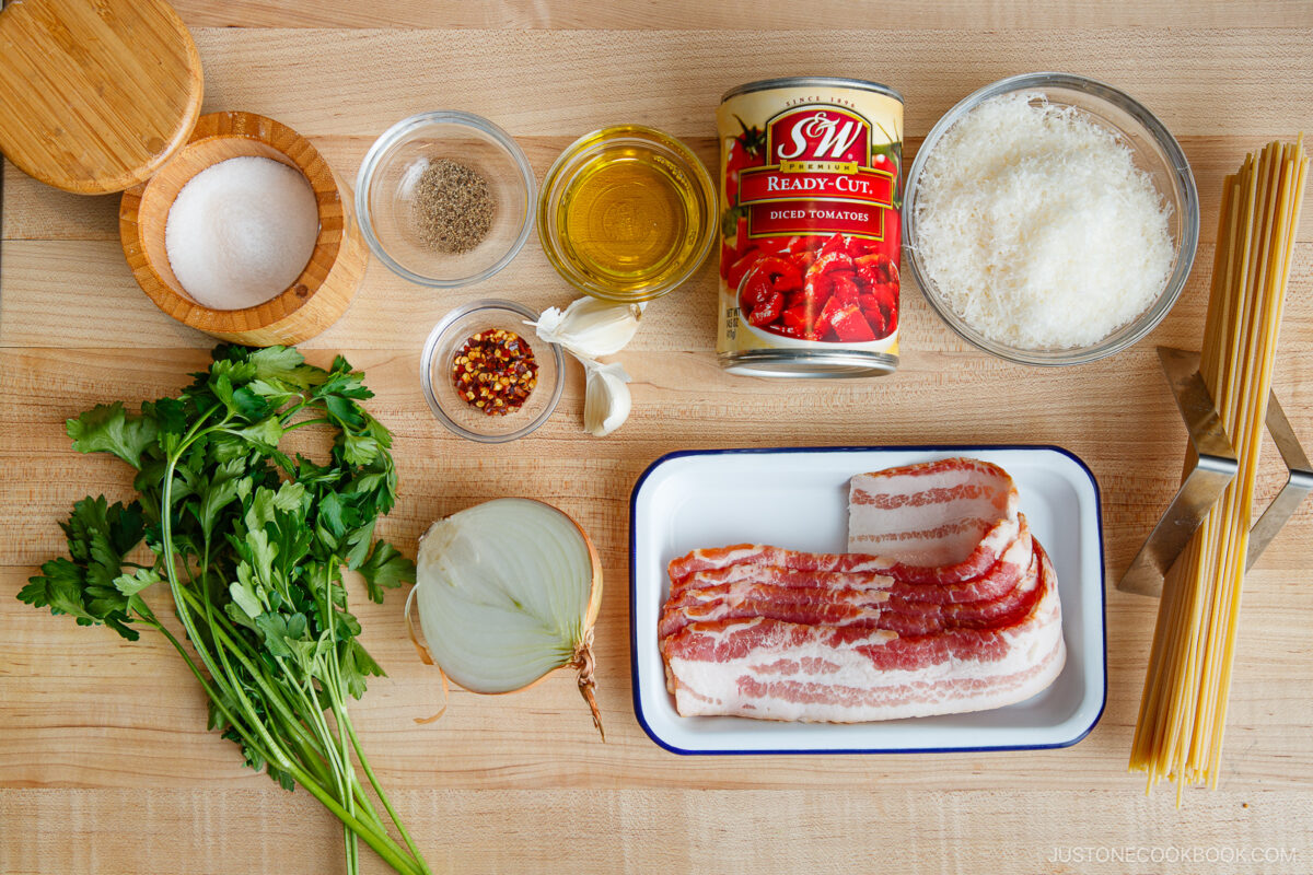 A wooden surface displays all the essentials for Tomato Bacon Pasta: bacon slices, dry spaghetti, canned diced tomatoes, grated cheese, olive oil, garlic cloves, parsley, onion, salt, pepper, and red pepper flakes in small bowls.
