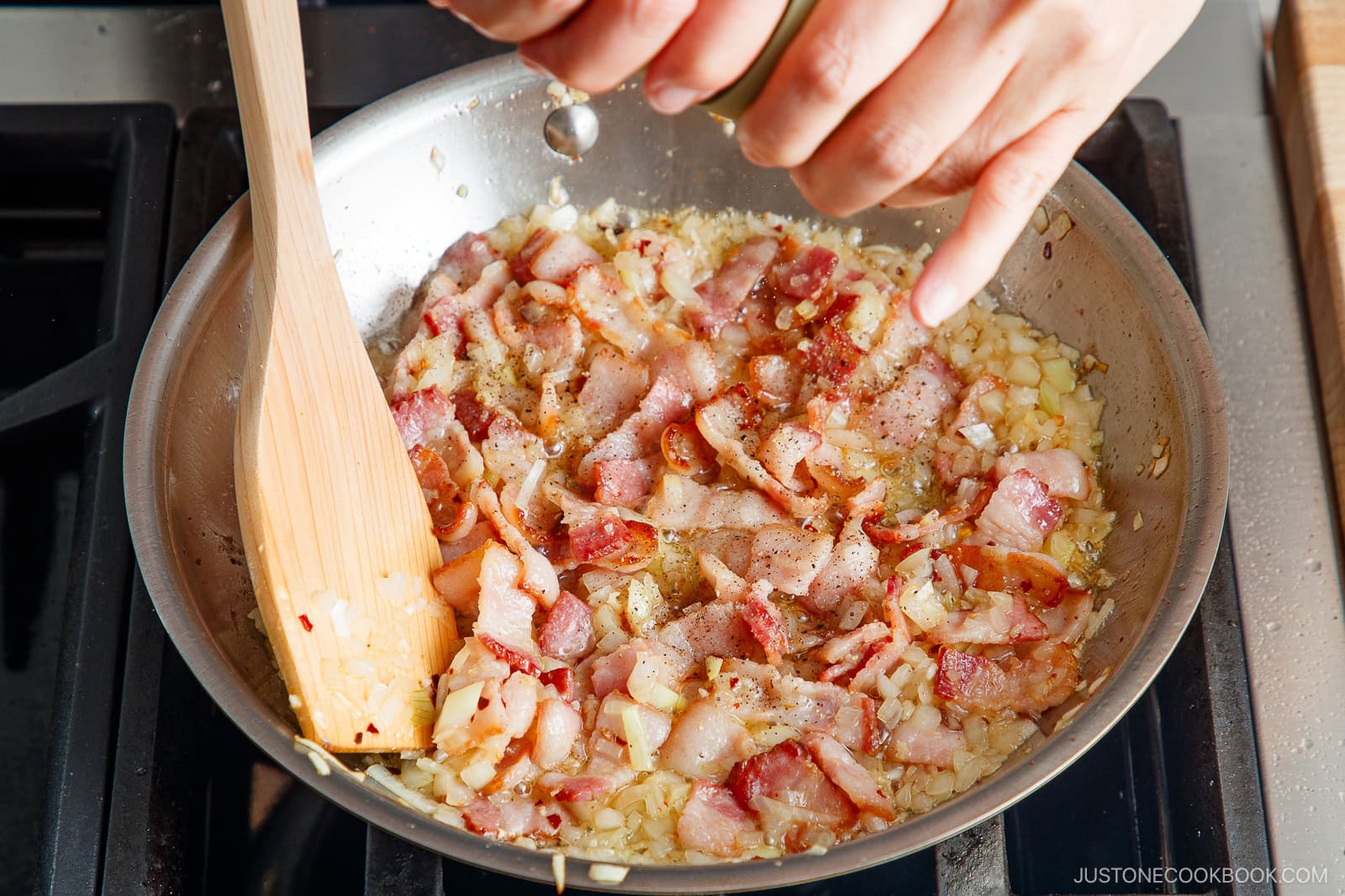 A hand seasons chopped bacon and onions cooking in a skillet, while a wooden spatula stirs the sizzling mixture—the savory base for Tomato Bacon Pasta—on a stovetop.