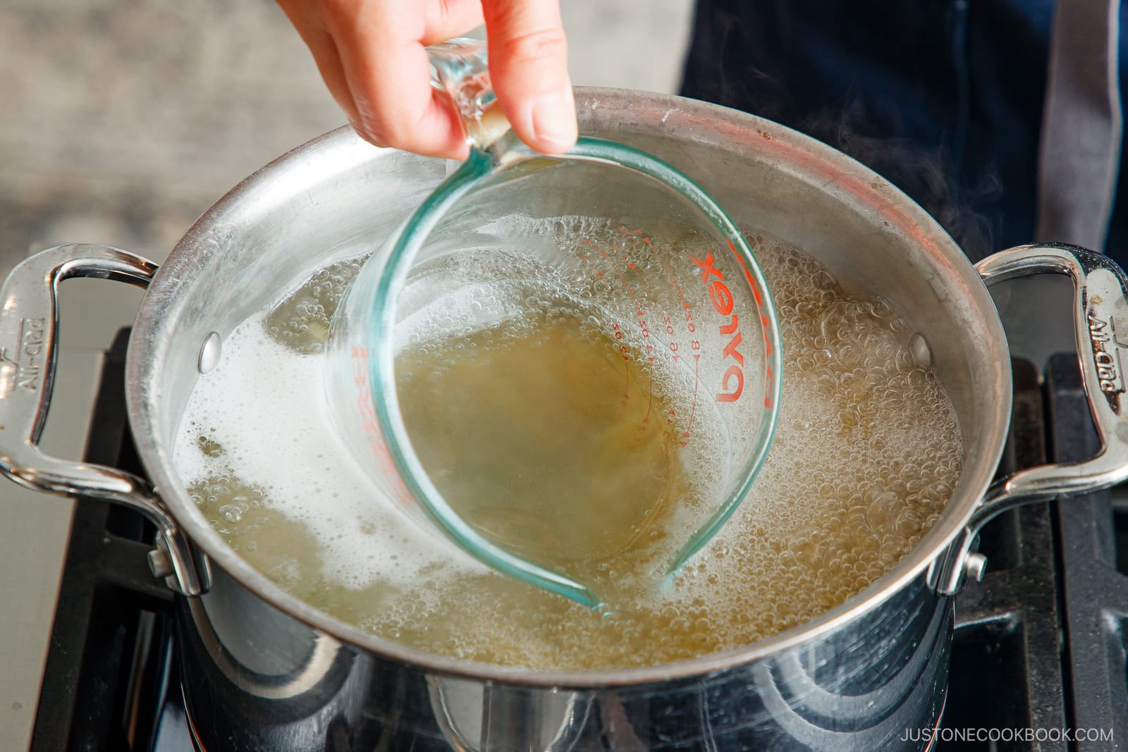 A hand holds a glass measuring cup, scooping hot, cloudy water from a pot of boiling noodles on a stovetop—perfect for making creamy Tomato Bacon Pasta.