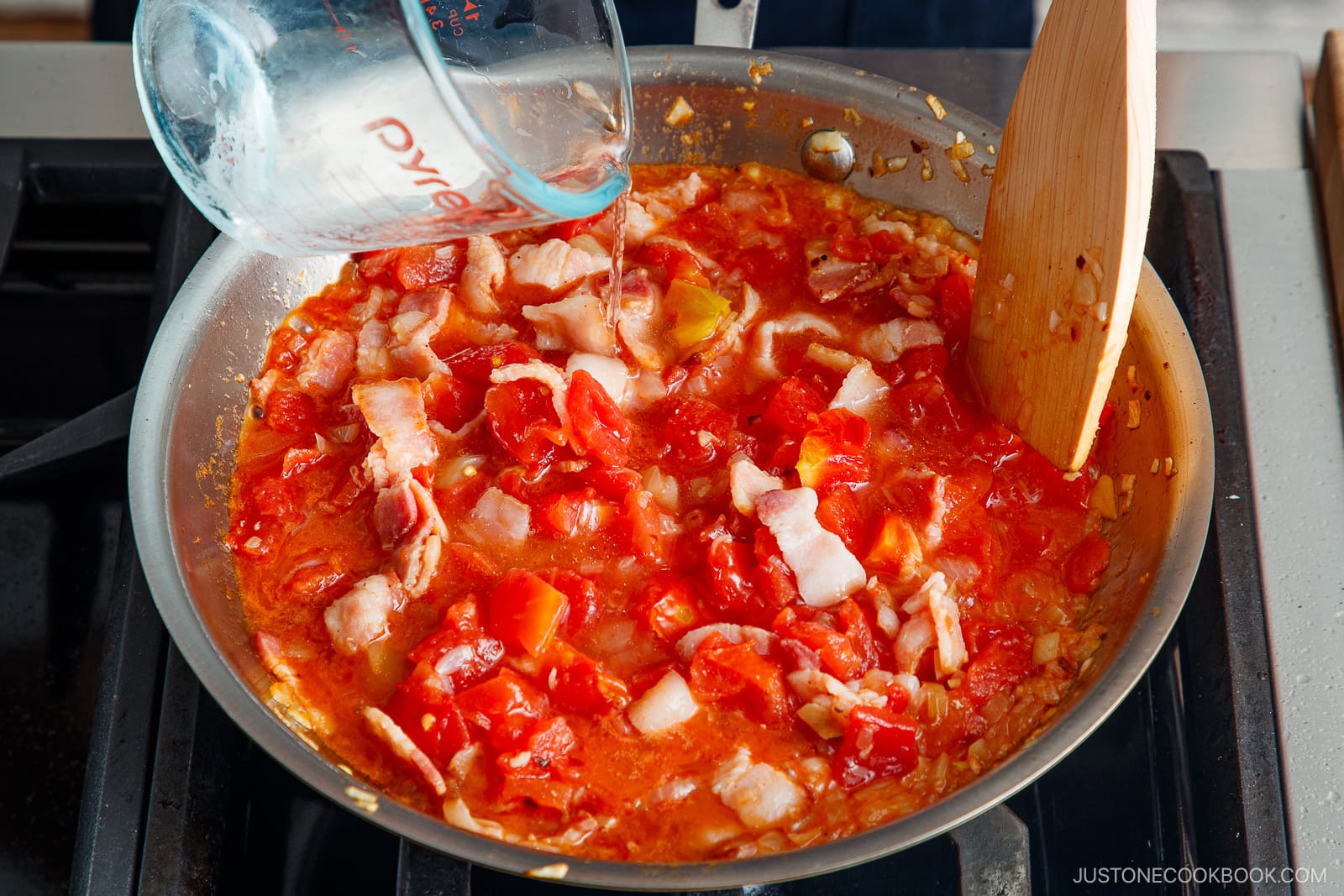 A hand pours water from a glass measuring cup into a skillet of simmering chopped tomatoes and bacon, stirring the Tomato Bacon Pasta mixture with a wooden spatula on a stovetop.