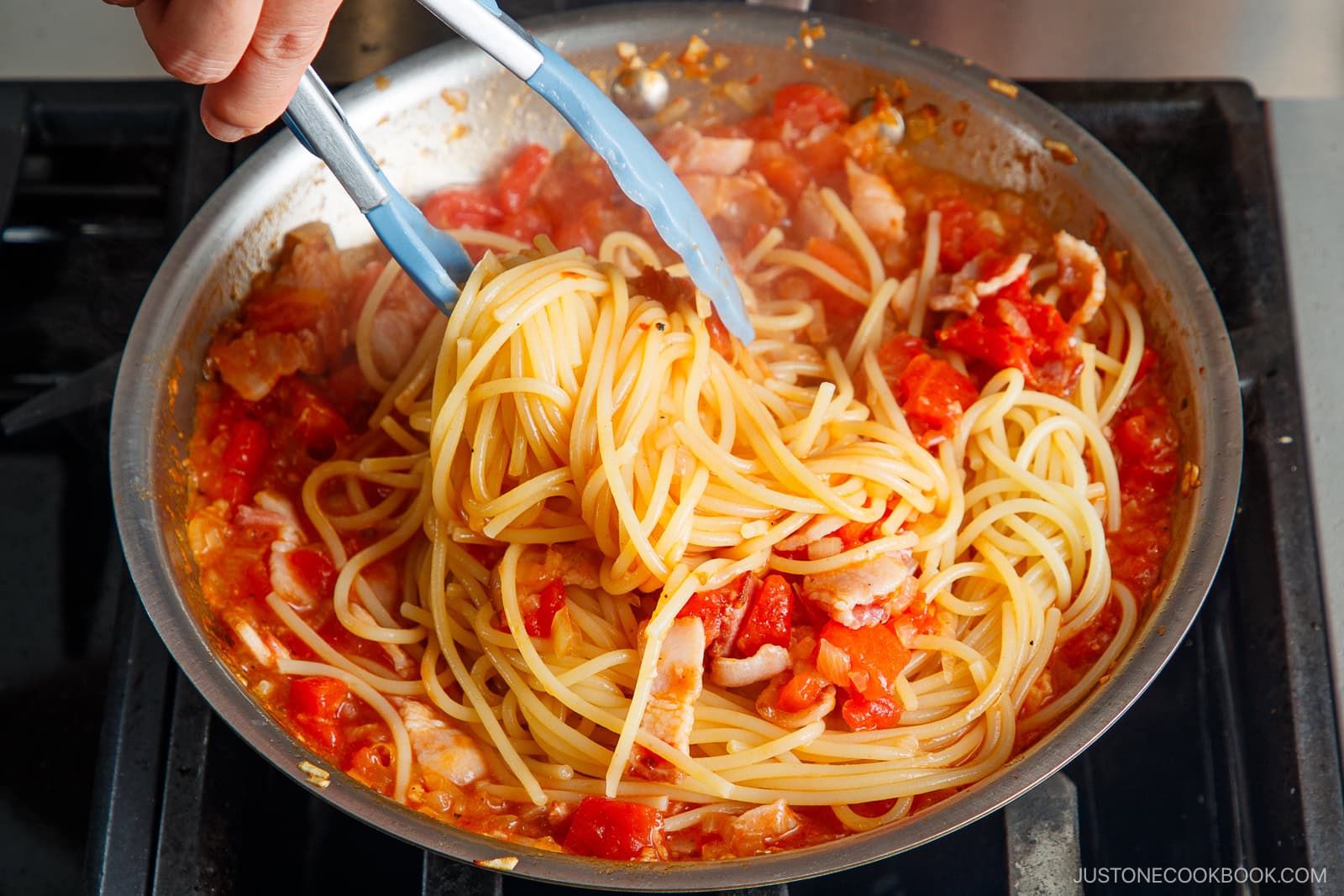 A hand uses tongs to mix Tomato Bacon Pasta with diced tomatoes and pieces of bacon in a skillet on a stovetop. The red, chunky sauce coats the spaghetti perfectly.