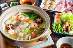A hot pot of chanko nabe (sumo stew) brims with shrimp, mushrooms, tofu, leafy greens, and vegetables, surrounded by plates of raw meat and dipping sauces—perfect for communal cooking and sharing at the table.