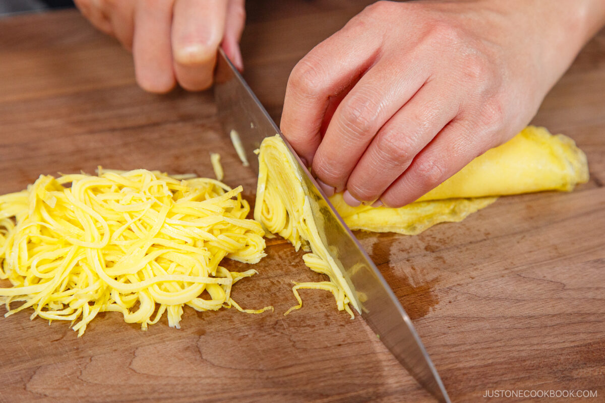 A person slices a rolled yellow omelet into thin strips on a wooden cutting board with a large chef’s knife—preparing the perfect topping for chirashi sushi. The sliced omelet pieces are neatly gathered beside the knife.