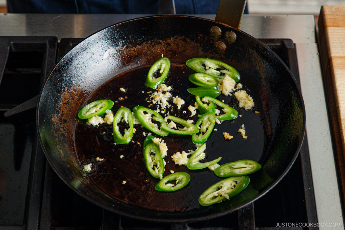 Sliced green chili peppers and minced garlic are being sautéed in a black skillet on a stovetop, forming the aromatic base for authentic gapao rice.