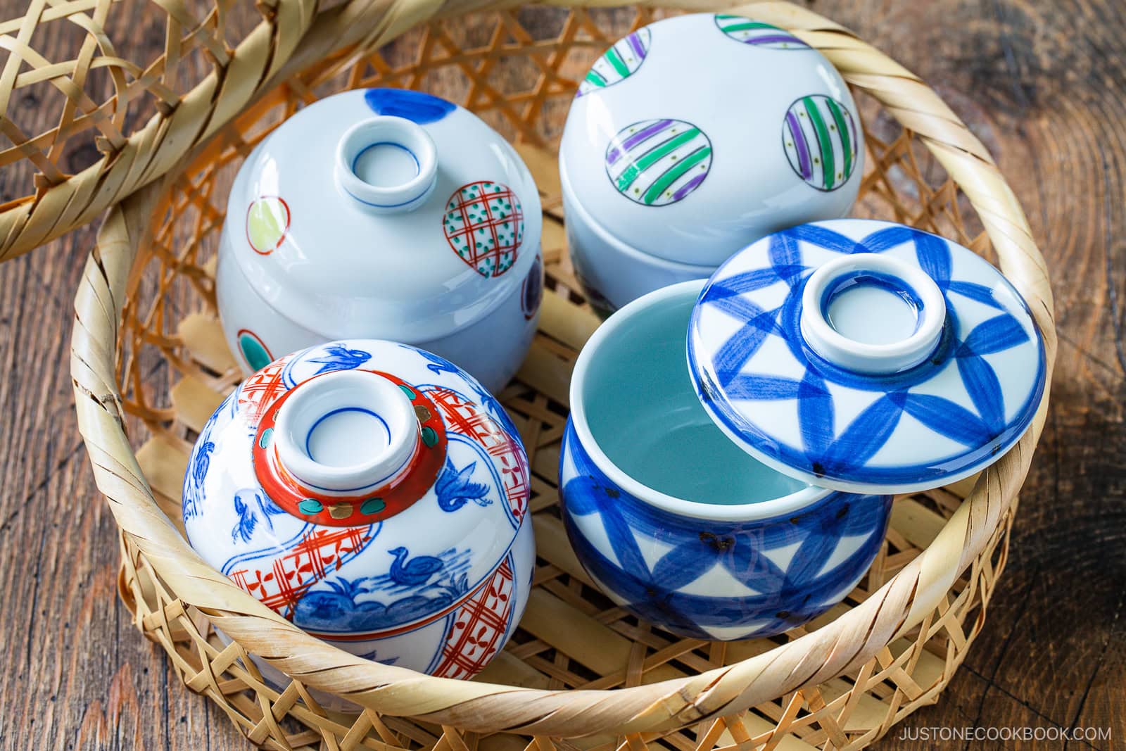 Three decorative Japanese ceramic bowls with lids, perfect for serving Quick & Easy Chirashi Sushi, feature colorful geometric and traditional patterns. They rest in a woven bamboo basket on a wooden surface, one lid slightly off to reveal the empty bowl inside.