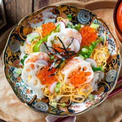 A decorative bowl of Quick & Easy Chirashi Sushi topped with shrimp, lotus root slices, cucumber, shredded egg, salmon roe, and nori strips, served on a wooden tray with red chopsticks and a cup of green tea.