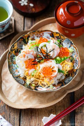 A colorful bowl of Quick & Easy Chirashi Sushi topped with ikura (salmon roe), sliced lotus root, shredded egg, cucumber, and seaweed sits on a wooden tray next to a cup of green tea and a miso soup bowl.