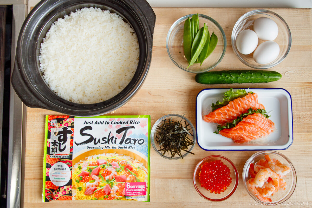 A wooden surface holds cooked rice in a pot, a cucumber, snap peas, eggs, raw salmon slices, shrimp, salmon roe, shredded seaweed, and a box of Sushi Taro seasoning mix—everything you need for Quick & Easy Chirashi Sushi.