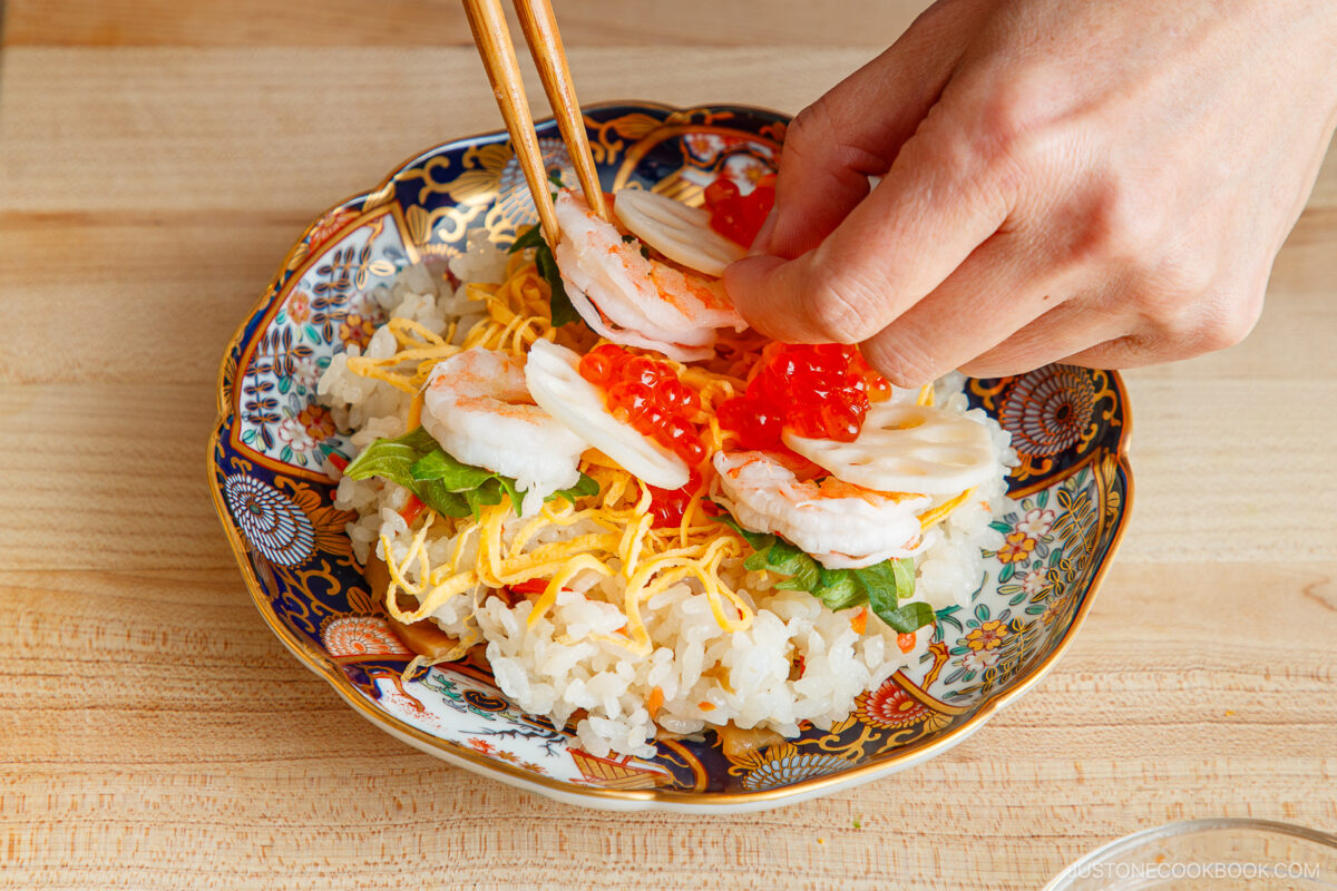 A hand uses chopsticks to place shrimp, sliced vegetables, and salmon roe on rice in a decorative bowl, assembling a colorful Quick & Easy Chirashi Sushi dish.