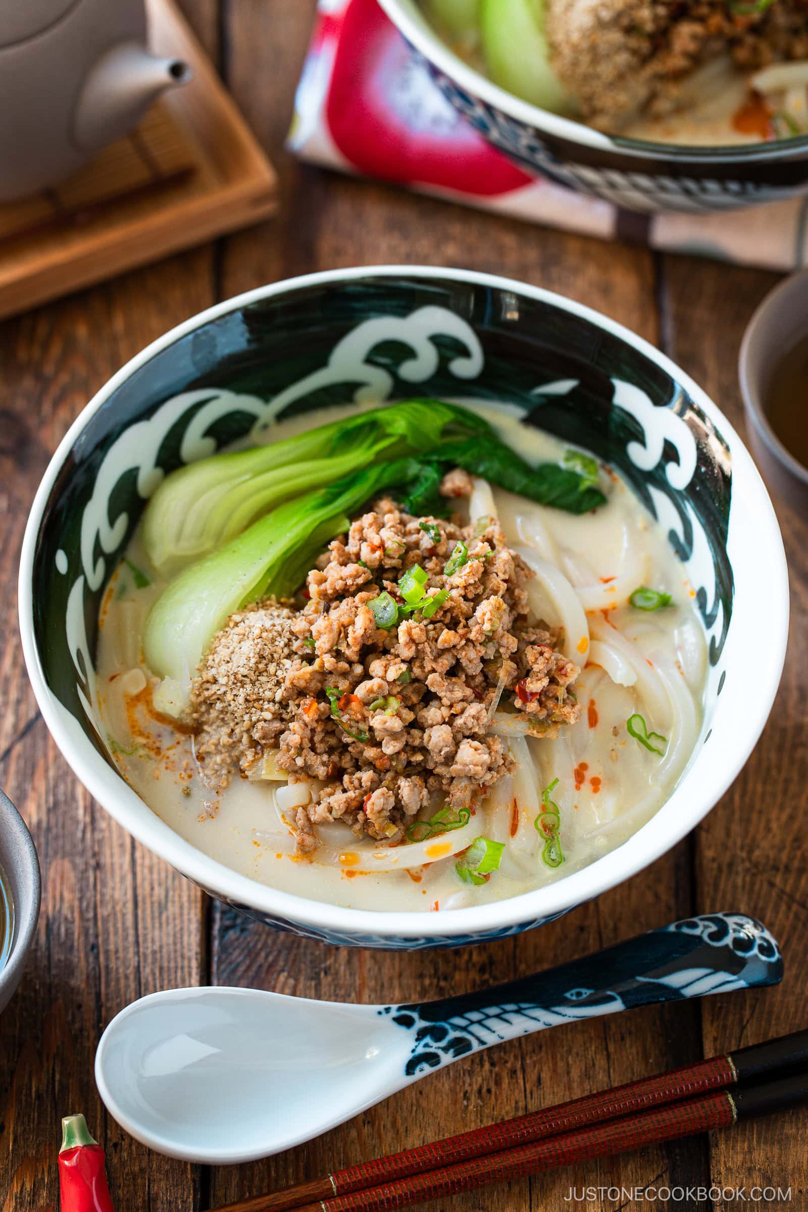 A bowl of tan tan udon noodle soup topped with ground meat, bok choy, sliced green onions, sesame seeds, and chili oil, served on a wooden table with a soup spoon and teapot nearby.