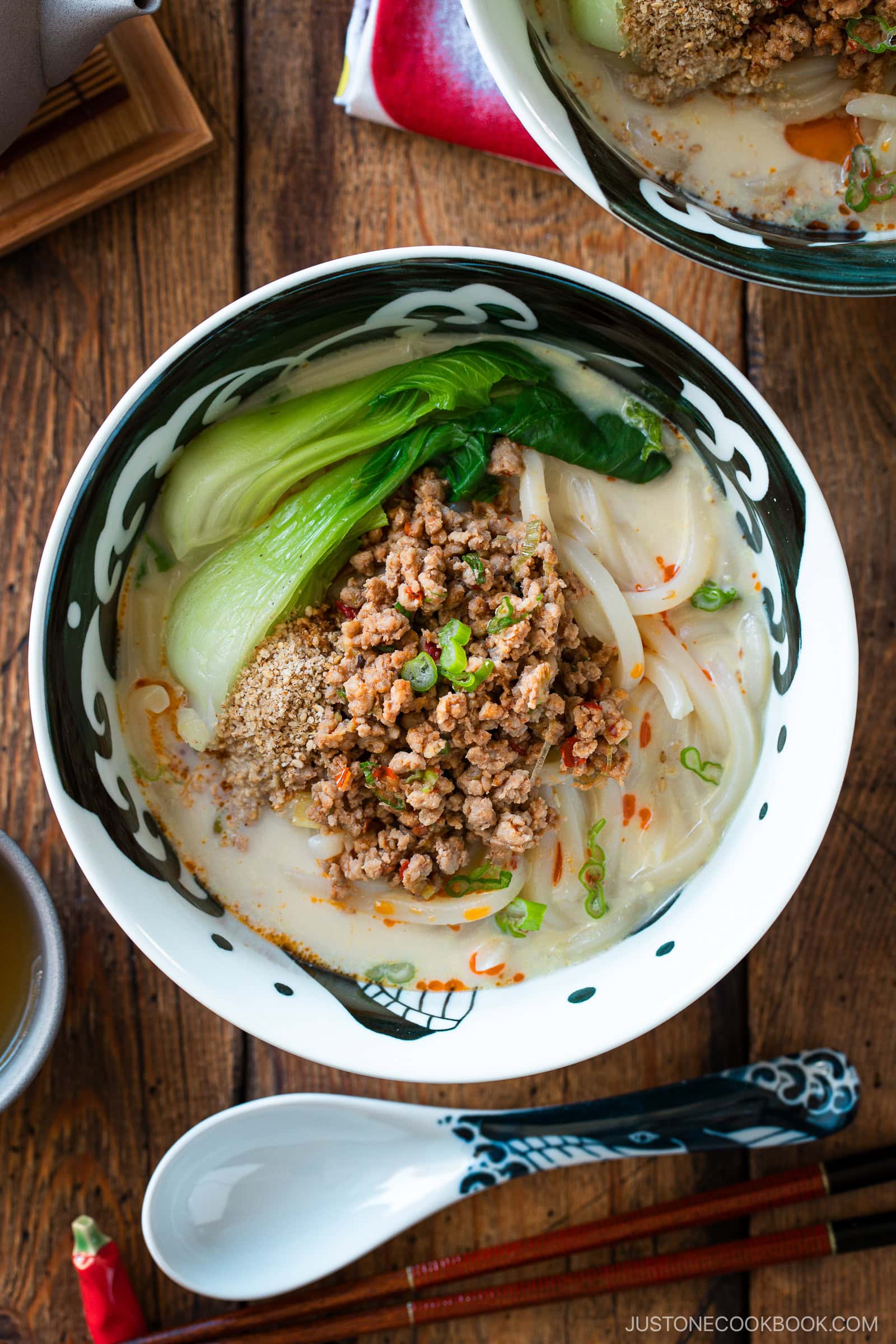 A bowl of tan tan udon noodle soup topped with ground meat, bok choy, green onions, and sesame seeds, served on a wooden table with a spoon, chopsticks, and another bowl partially visible.
