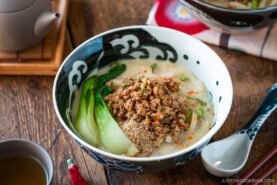 A bowl of creamy tan tan udon broth ramen topped with ground meat, bok choy, sesame seeds, and green onions sits on a wooden table with a spoon, chopsticks, teacup, and teapot nearby.