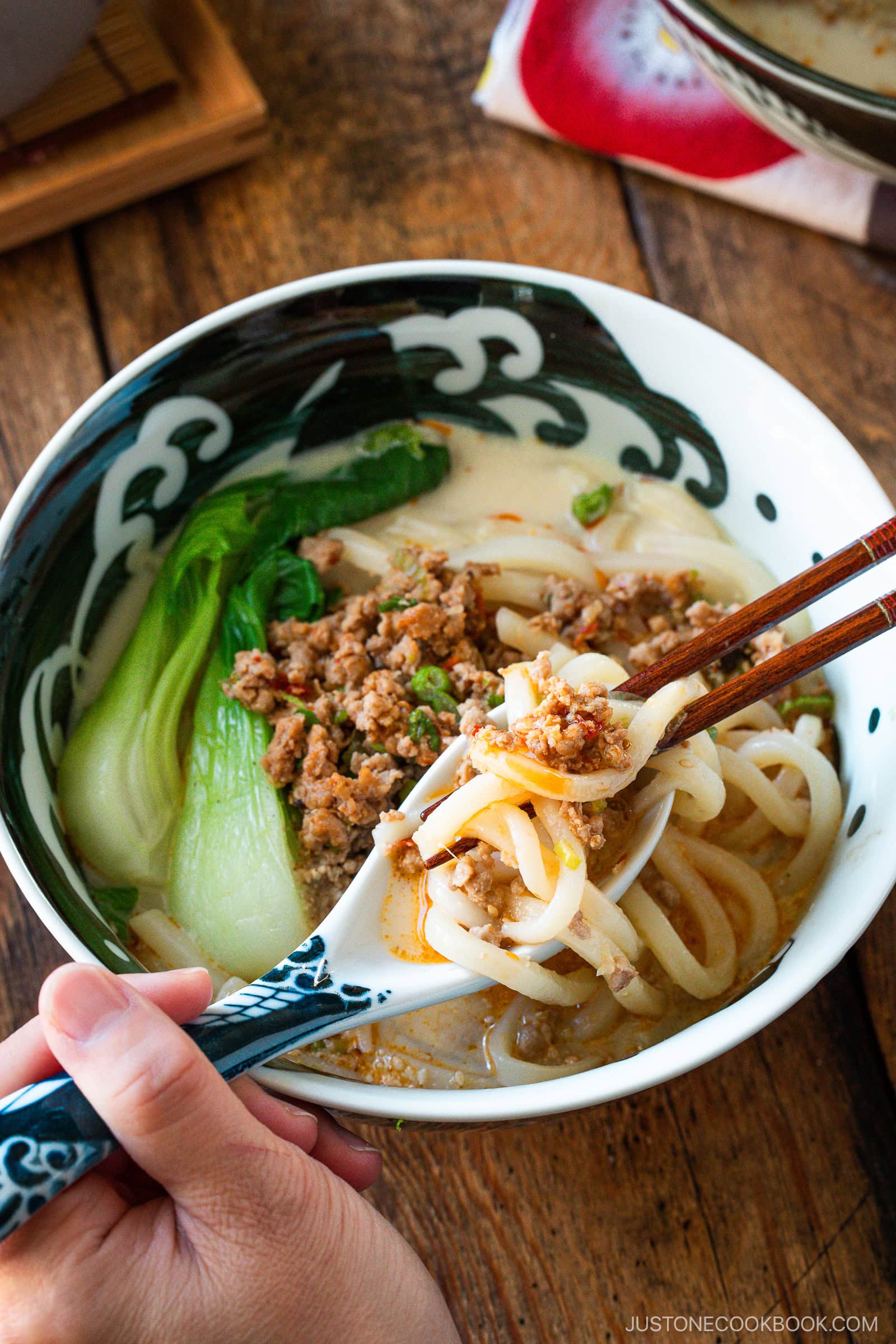 A hand using chopsticks lifts tan tan udon noodles from a bowl of soup with ground meat, bok choy, and a creamy broth on a wooden table.