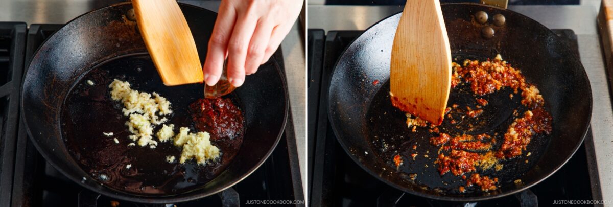 A hand uses a wooden spatula to stir minced garlic and red chili paste in a black skillet on a stove, mixing and sautéing the base for flavorful tan tan udon as the ingredients begin to cook.