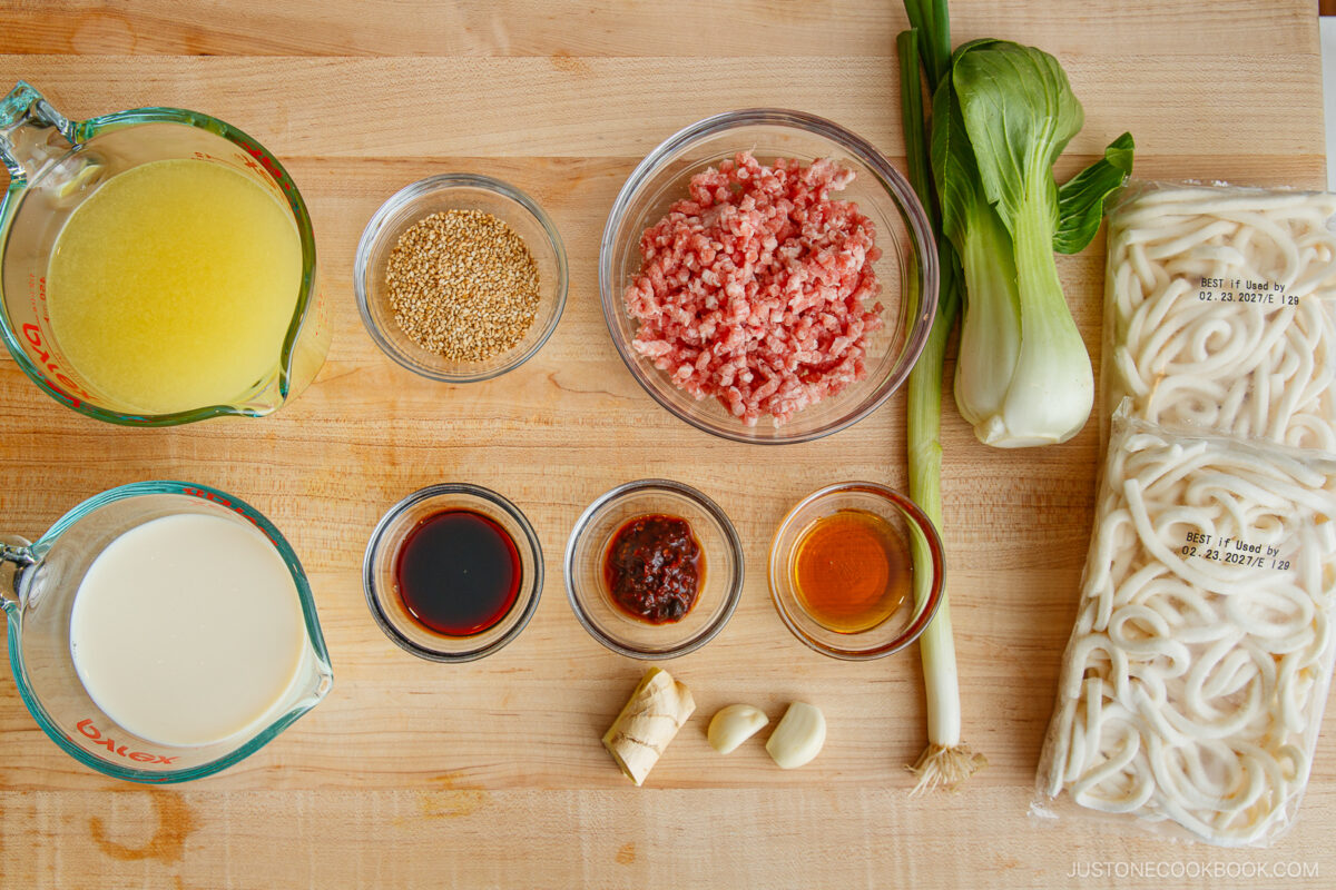 Overhead view of tan tan udon ingredients on a wooden surface: chicken broth, sesame seeds, ground pork, bok choy, udon noodles, milk, soy sauce, chili paste, sesame oil, ginger, garlic, and green onion.