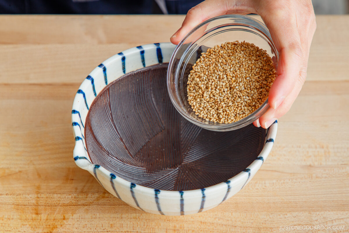 A hand holds a small glass bowl of sesame seeds above a ceramic bowl with a ridged interior on a wooden surface, preparing to add them as part of tan tan udon preparation.