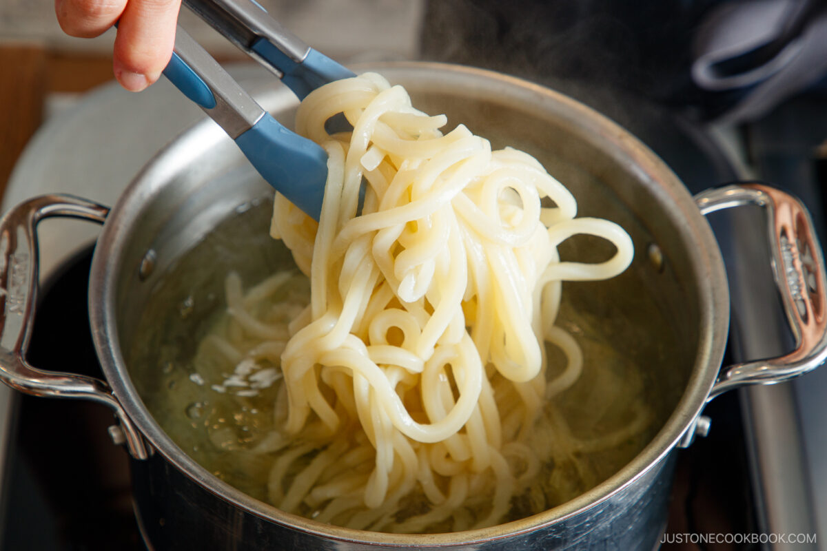 A hand uses tongs to lift cooked tan tan udon noodles from a pot of boiling water on a stovetop.