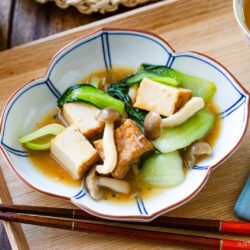 A bowl of bok choy and tofu stir-fry with mushrooms and savory sauce is served on a wooden tray with chopsticks and a cup of tea, beautifully presented in a white and blue flower-shaped bowl.